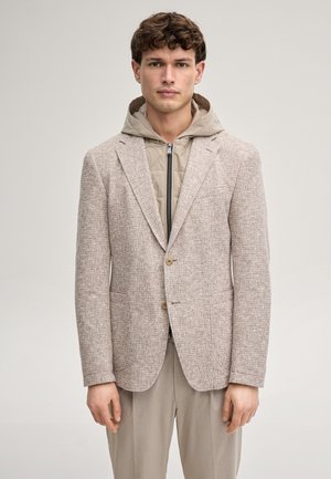 Junger Mann, der einen hellbeigen, strukturierten Blazer über einem taupefarbenen, gesteppter Reißverschluss-Kapuzenpullover und passenden beigen Hosen trägt, stehend vor einem einfarbigen Hintergrund.