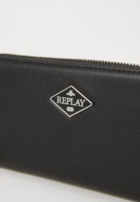 Crni kožni novčanik s metalnim dijamantskim izrezom na kojem stoji "REPLAY" i "1981", glatke teksture i zatvaranjem na zip.