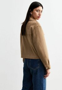 Giacca cropped in suede marrone con una texture liscia, maniche lunghe e dettaglio con bottoni. I jeans in denim blu a vita alta completano il look.