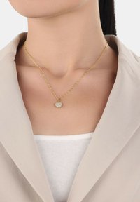 Collana in tono oro con catena, caratterizzata da un pendente a forma di cuore decorato con cristalli trasparenti, indossata su una maglietta bianca e sotto un giacca beige.