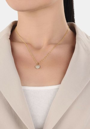 Collar de cadena en tono dorado con un colgante en forma de corazón adornado con cristales transparentes, que se lleva sobre una blusa blanca y debajo de una chaqueta beige.