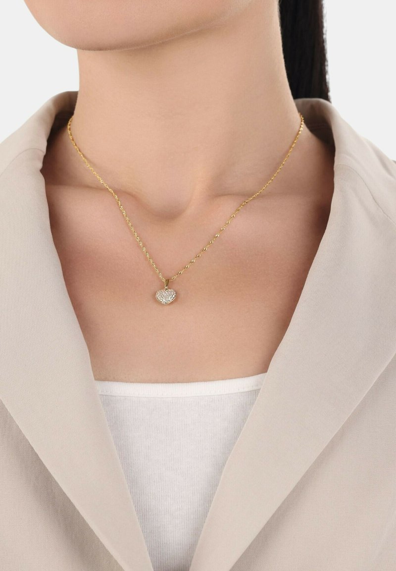 Collana in tono oro con catena, caratterizzata da un pendente a forma di cuore decorato con cristalli trasparenti, indossata su una maglietta bianca e sotto un giacca beige.