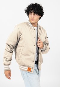 Beżowa kurtka bomberka z prążkowanymi mankietami i dolnym wykończeniem, z zapięciem na zatrzaski, przednimi kieszeniami i pomarańczową metką. Nosi ją osoba w białej koszulce.