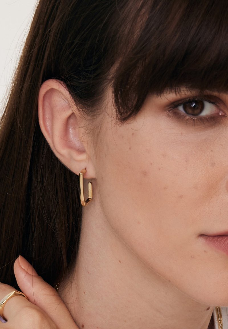 Złoty geometryczny kolczyk w kształcie obręczy o czystym, kanciastym designie. Polerowane wykończenie podkreśla elegancki kontur, dopełniając ucho noszącego.