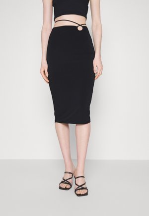 Mujer que lleva una falda lápiz negra de talle alto con un detalle de anillo en la cintura, combinada con un top corto negro y sandalias negras con tiras.