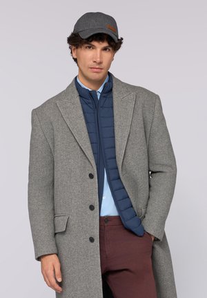 Cappotto grigio a spina di pesce sopra una giacca imbottita blu, camicia azzurra, pantaloni bordeaux e cappello grigio con dettaglio in pelle marrone.