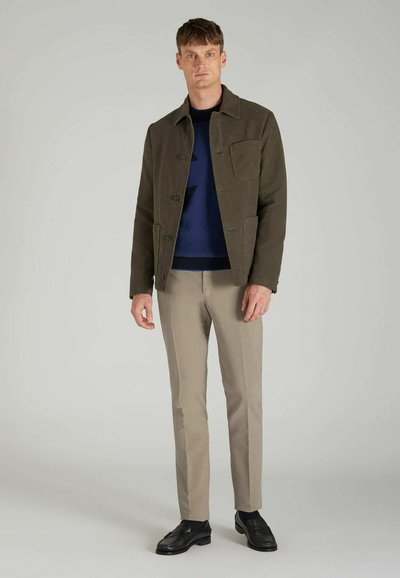 Olive green jacket with a collared design, front pockets, worn over a navy blue sweater, paired with beige trousers and black shoes.