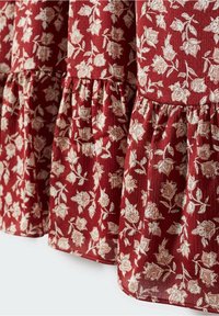 Der rote Stoff mit floralem Muster hat eine strukturierte Oberfläche mit cremefarbigen Blumenmustern und einem gerafften Saum. Verstellbare Passform möglich.