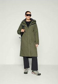 Donna con un lungo cappotto verde oliva, pantaloni neri, scarpe da trekking grigie, occhiali da sole neri e un zaino nero, su uno sfondo semplice.
