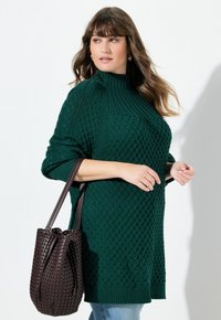 Maglione lavorato a maglia verde smeraldo con motivi testurizzati e collo alto, abbinato a una borsa a secchiello intrecciata di colore marrone scuro.