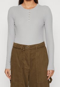 Top a maniche lunghe ribattuto grigio chiaro con scollatura rotonda e patta con sette bottoni, abbinato a pantaloni cargo marroni a vita alta con cintura.