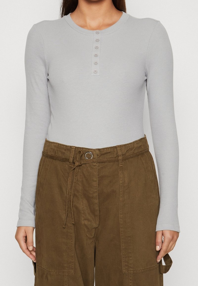 Top a maniche lunghe ribattuto grigio chiaro con scollatura rotonda e patta con sette bottoni, abbinato a pantaloni cargo marroni a vita alta con cintura.