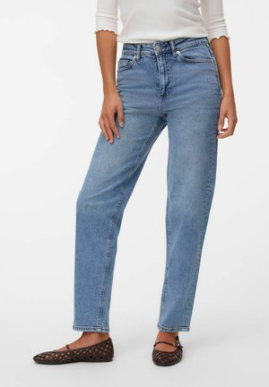 Vrouw draagt lichtblauwe, rechte spijkerbroek met hoge taille, witte top met lange mouwen en donkerbruine gevlochten platte schoenen, staand met ontspannen handen.