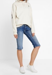 Person som bär en krämfärgad Puma-hoodie, knälånga blå denimshorts och vita sneakers, stående mot en vit bakgrund.