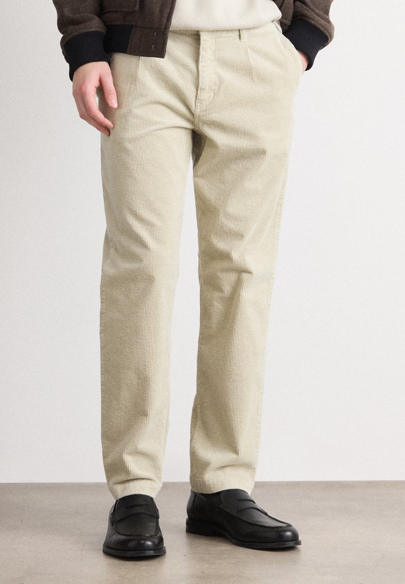 Calças de veludo cotelê bege com uma superfície texturizada, corte slim, e bolsos standard, combinadas com sapatos mocassim pretos num chão liso.