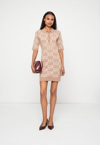 Beżowa sukienka z kołnierzykiem i krótkimi rękawami, zdobiona wzorem przeplatających się logo. W komplecie burgundowa torebka i szpilki.