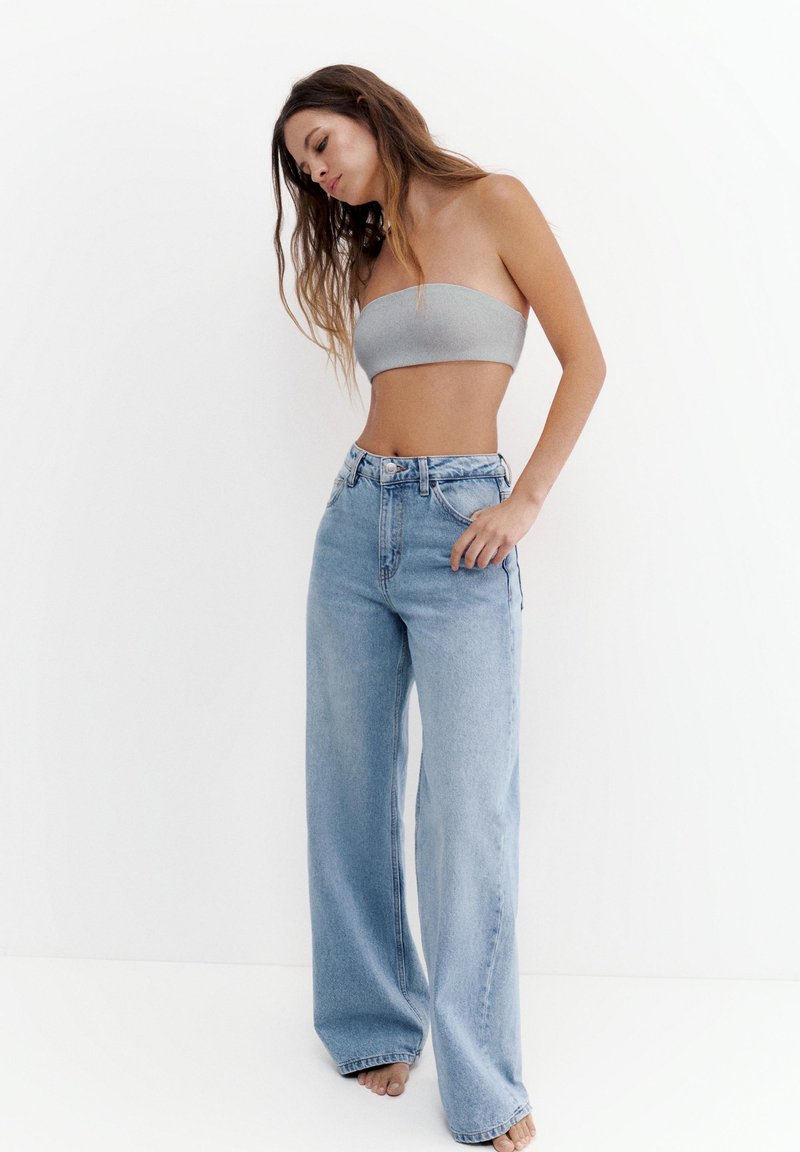 Femme aux longs cheveux portant un haut tube gris et un jean bleu taille haute à jambes larges, debout pieds nus devant un fond blanc.