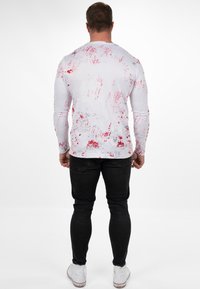 Wit overhemd met lange mouwen en rode spetterpatronen, gecombineerd met zwarte jeans en witte sneakers. De achterkant toont de grafische textuur en het ontwerp.
