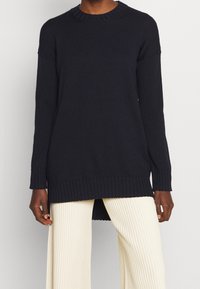 Marineblau gerippter Strickpullover mit langen Ärmeln, Rundhalsausschnitt und Seitenschlitzen am Saum, kombiniert mit cremefarbenen, plissierten Hosen.