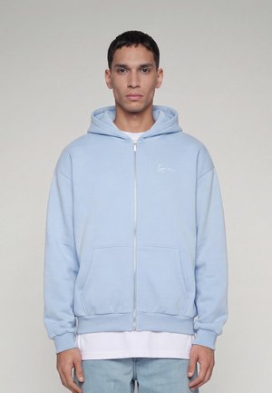 Jeune homme portant un sweat à capuche zippé bleu clair avec des poches, une chemise blanche en dessous et un jean bleu clair, debout devant un fond uni.
