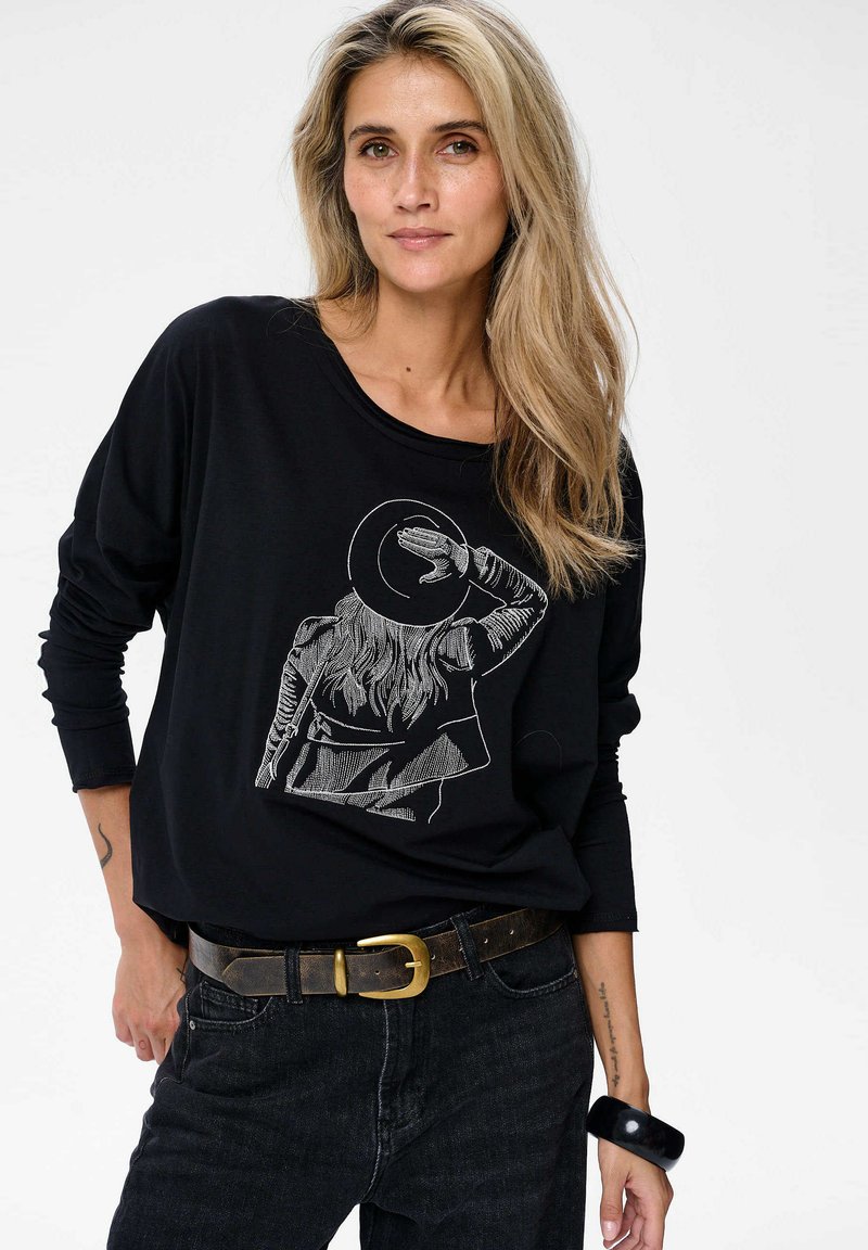 Zwarte longsleeve tshirt met een afbeelding van een persoon in een hoed, gecombineerd met donkere jeans en een bruine riem met een gouden gesp.