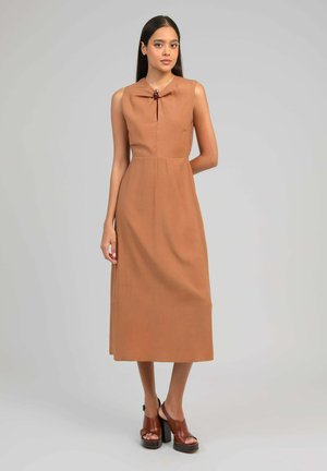 Jeune femme debout portant une robe mi-longue marron sans manches avec une découpe en forme de clé sur le devant et des talons compensés marron sur fond gris.