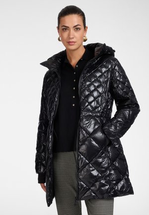 Donna che indossa un cappotto trapuntato nero lucido con cappuccio, camicia nera con bottoni, pantaloni a quadri e orecchini a cerchio dorati su sfondo semplice.