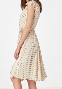 Krämfärgad klänning med bruna prickar, korta volangärmar, åtsittande midja och en flödande kjol, tillverkad av lätt, strukturerad tyg.