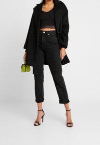 Frau trägt schwarzen, kurzen Spitzenoberteil, schwarzen strukturierten Mantel, ausgefranste schwarze Jeans, schwarze High Heel Sandalen und hält eine kleine grüne Handtasche.