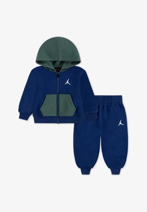 Set di felpa e pantaloni blu navy con cappuccio verde, zip frontale, tasca a marsupio e accenti bianchi del logo sul petto e sulla gamba dei pantaloni.