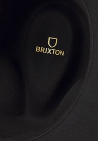 Black felt hat featuring a smooth texture and a rounded crown. "Brixton" logo in gold is prominently displayed inside the brim.