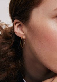 Złoty kolczyk w kształcie obręczy zaprezentowany na uchu, charakteryzujący się gładkim, wypolerowanym wykończeniem i okrągłym kształtem, częściowo osadzony na falującymi brązowymi włosami.