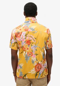 Chemise à manches courtes avec un fond jaune et des motifs floraux vibrants en rose, rouge et bleu. Tissu léger avec une coupe décontractée.
