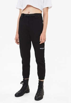 Person iført sorte cargobukser med bælte og elastiske manchetter, sorte snørebåndsstøvler og en kort hvid t-shirt, stående mod en hvid baggrund.