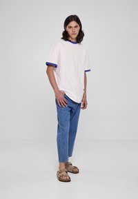 Lys rosa T-skjorte med blå kontrastdetaljer på rund hals og ermer, kombinert med avslappede blå denim jeans og brune sandaler.