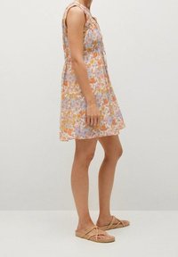 Robe fleurie dans des couleurs pastel avec un design sans manches, une coupe au genou et un tissu léger. Associée à des sandales brunes plates et à lanières.