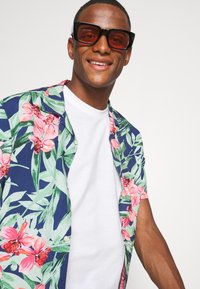 Kurzarmhemd mit navy Hintergrund und großen rosa und grünen Blumenmustern; getragen über einem weißen Rundhals-T-Shirt.