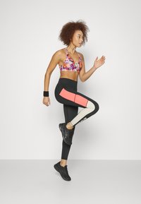 Nainen kukkakuosisessa urheiluliivissä ja värilohkoleggingsit hyppii paikoillaan yksinkertaista valkoista taustaa vasten.