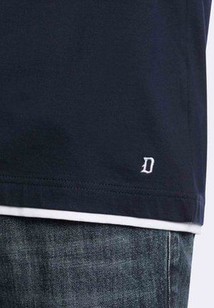 Orlo di camicia blu navy scuro con piccolo logo bianco ricamato a forma di "D" sopra la vita dei jeans in denim blu.
