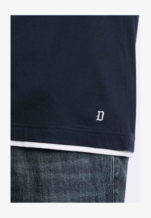 Orlo di camicia blu navy scuro con piccolo logo bianco ricamato a forma di "D" sopra la vita dei jeans in denim blu.