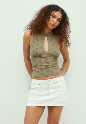 Młoda kobieta o kręconych włosach, ubrana w zielony, kwiatowy top bez rękawów z dekoltem typu keyhole oraz białą mini spódniczkę, stojąca na jasnym tle.