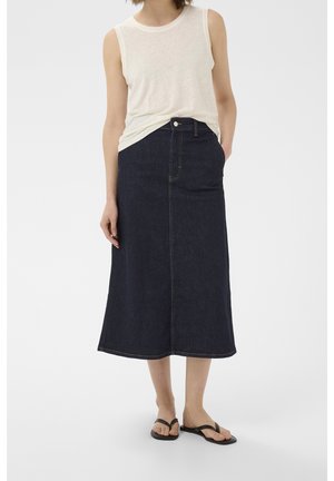 Femme portant un haut blanc sans manches, une jupe midi en jean bleu foncé et des sandales noires, debout avec une main dans la poche de la jupe.