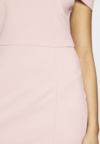 Torso et bras d'une femme portant une robe ajustée rose clair avec des coutures visibles et une texture de tissu lisse sur fond blanc.