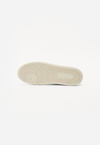 Suola della scarpa vista dal basso, realizzata in gomma di colore beige chiaro. Presenta un motivo a zig-zag e un logo circolare sul tallone.
