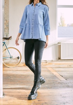 Vrouw draagt een lichtblauw denimtop met knopen, zwarte skinny jeans en zwarte veterschoenen, staand binnen bij een vintage fiets.