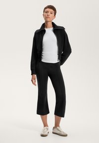 Veste noire courte avec col montant, assortie à un haut blanc ajusté et à des leggings noirs évasés. Le modèle porte des baskets légères. Textures de tissu lisses.