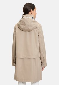 Veste imperméable beige avec capuche, manches longues et poignets à boutons-pression. Présente un design en couches avec un ourlet arrière plus long.