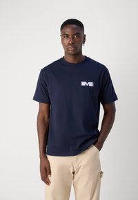 Námořnická modrá bavlněná tričko s krátkými rukávy, kulatým výstřihem a bílým logem na levé části hrudi, v kombinaci se světle béžovými kalhotami.