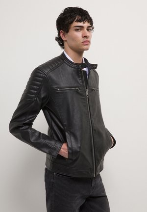 Jeune homme portant une veste en cuir noir matelassée avec des poches zippées sur la poitrine et les mains dans les poches latérales, se tenant contre un mur uni.