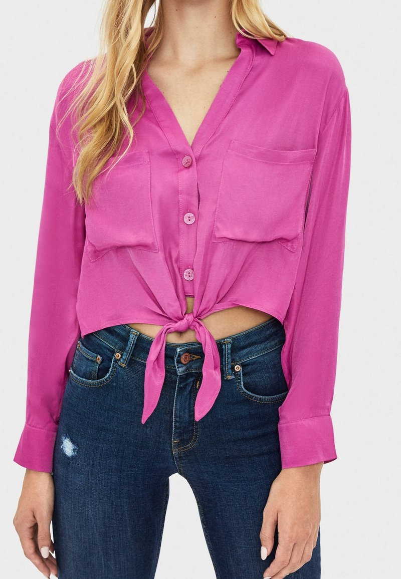 Blusa a maniche lunghe fucsia con un nodo sul davanti, caratterizzata da due tasche sul petto e una texture satinata liscia, abbinata a jeans in denim scuro.
